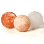 Three handcrafted smooth round Himalayan pink salt massage balls in red, peach, and white crystal shades.