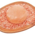 Handcrafted smooth round Himalayan pink salt massage stone resting on coarse pink rock salt crystals within a wooden dish