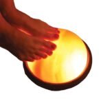 Bare feet resting on an illuminated handcrafted Himalayan pink salt foot detox dome lamp mounted on a dark round wooden base