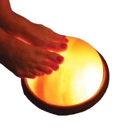 Bare feet resting on an illuminated handcrafted Himalayan pink salt foot detox dome lamp mounted on a dark round wooden base